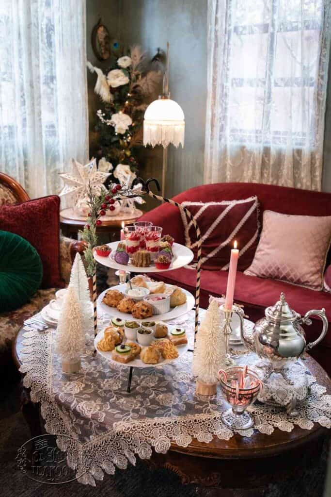 A tiered tray with pastries and desserts sits on a lace-covered table, surrounded by white decorative trees, a lit pink candle, a silver teapot, and velvet sofas in a cozy, elegant room decorated for the holidays.