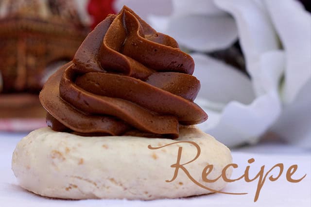 A cookie topped with a swirl of rich chocolate frosting sits on a white surface, with the word “Recipe” written in cursive in the foreground. A soft, blurred background features a white flower.
