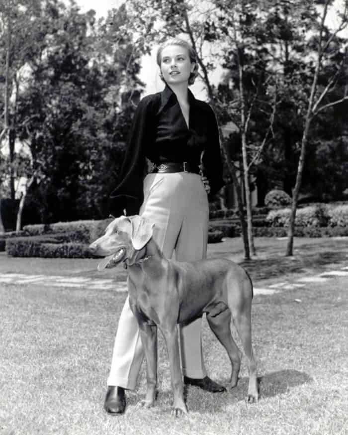 A woman in a black blouse and light trousers stands outdoors on grass with a large dog at her side. Trees and bushes are visible in the background. The image is in black and white.