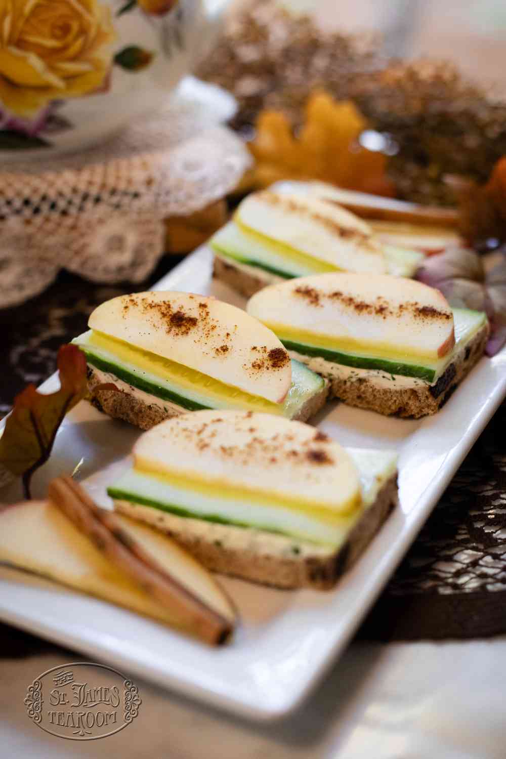 Three open-faced sandwiches topped with apple slices, cucumber, and cinnamon are arranged on a rectangular white plate, with cinnamon sticks and apple slices beside them. Inspired by an Apple Boursin Tea Sandwich recipe, a floral teacup is partially visible in the background.