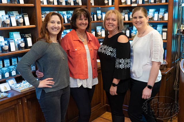 Four women stand side by side, smiling, in front of wooden shelves stocked with packages inside a shop or store.