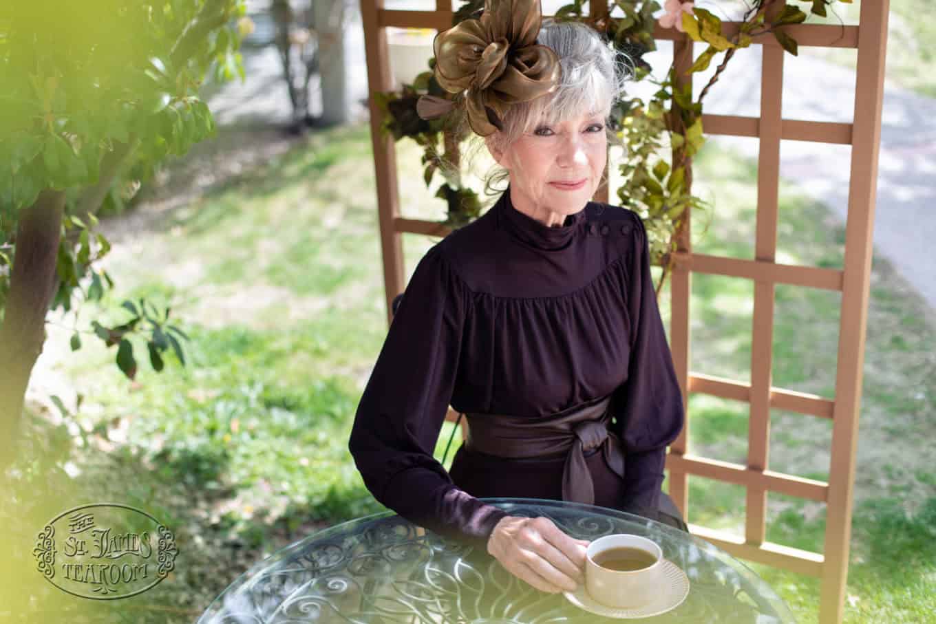 An elderly woman with gray hair and a large brown flower hat sits at a glass table outdoors, holding a cup of coffee and smiling gently. She wears a dark dress and greenery surrounds the scene.