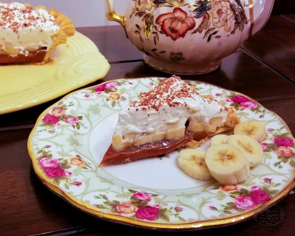 A slice of classic banoffee pie topped with whipped cream and chocolate shavings sits on a floral plate, next to fresh banana slices. A teapot and another delicious banoffee pie are in the background.