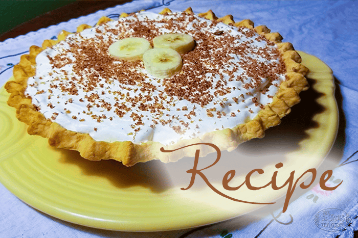 Banoffee pie with whipped topping and chocolate shavings, garnished with banana slices, sits on a yellow plate. The word “Recipe” is written in cursive across the bottom right corner.