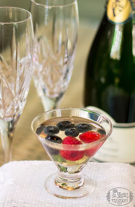 A small glass filled with clear liquid, blueberries, and raspberries sits on a white cloth—an elegant presentation reminiscent of Berry Champagne Gelées. Crystal glasses and a bottle are in the blurred background, completing the sophisticated scene.
