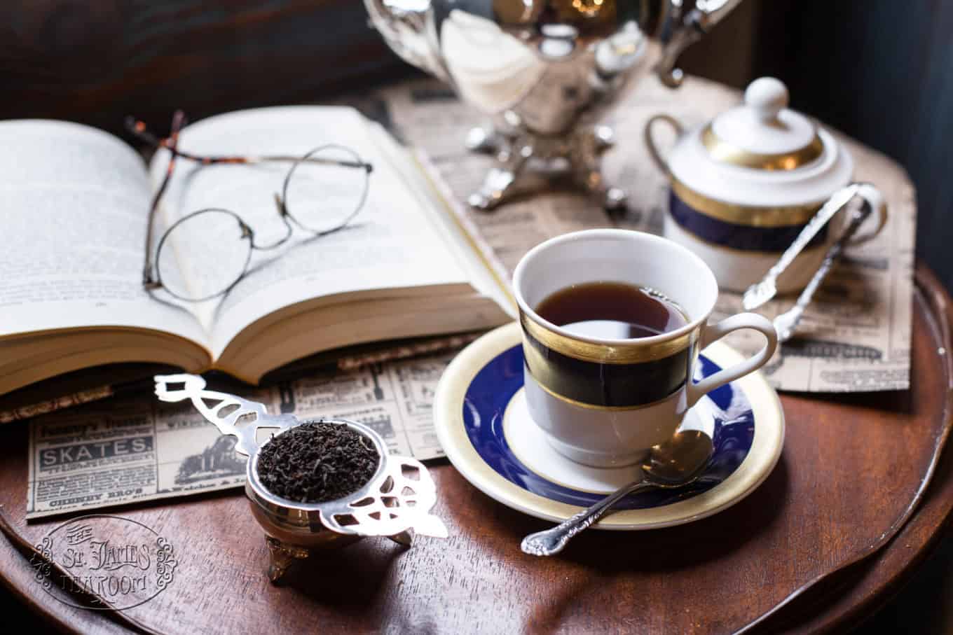 A cup of tea on a saucer sits beside an open book with glasses, a tea strainer filled with loose tea, a sugar bowl, and newspapers on a wooden table.