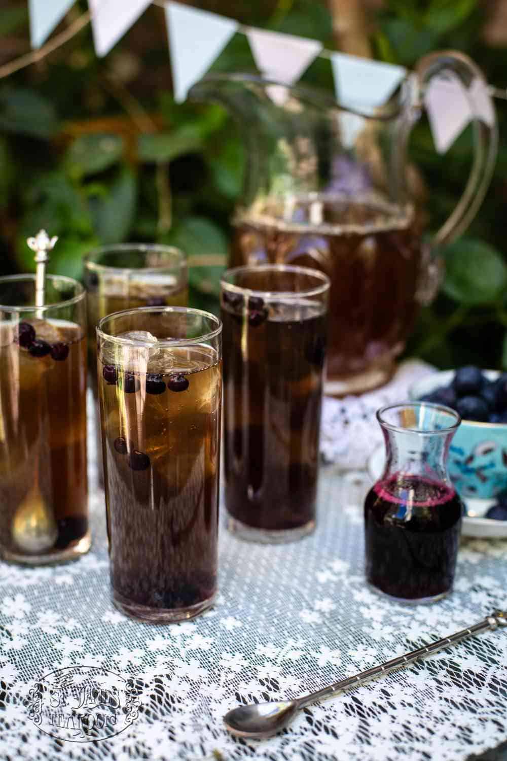 Four tall glasses of iced tea with blueberries rest on a lace tablecloth, showcasing a refreshing Iced Tea Recipe. A pitcher of tea, a small jar of blueberry syrup, a bowl of blueberries, and blue-and-white bunting in the background complete this charming summer scene.