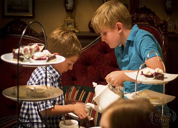 Two young boys are having tea together at a table set with desserts and treats on tiered trays. One boy pours tea while the other holds a cup. The setting looks cozy and elegant.