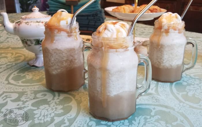 Three mason jar mugs filled with frothy iced coffee, reminiscent of Butterbeer Ice Cream Floats, are topped with whipped cream and caramel sauce. Each is finished with a metal straw and sits on a patterned tablecloth next to a teapot and a tray of pastries.