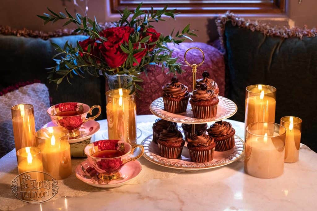 A cozy table setting with chocolate cupcakes on a tiered stand, pink teacups filled with tea, several lit candles, and a vase of red roses creates a warm, inviting atmosphere—perfect for sharing your favorite Iced Tea Recipe with friends.
