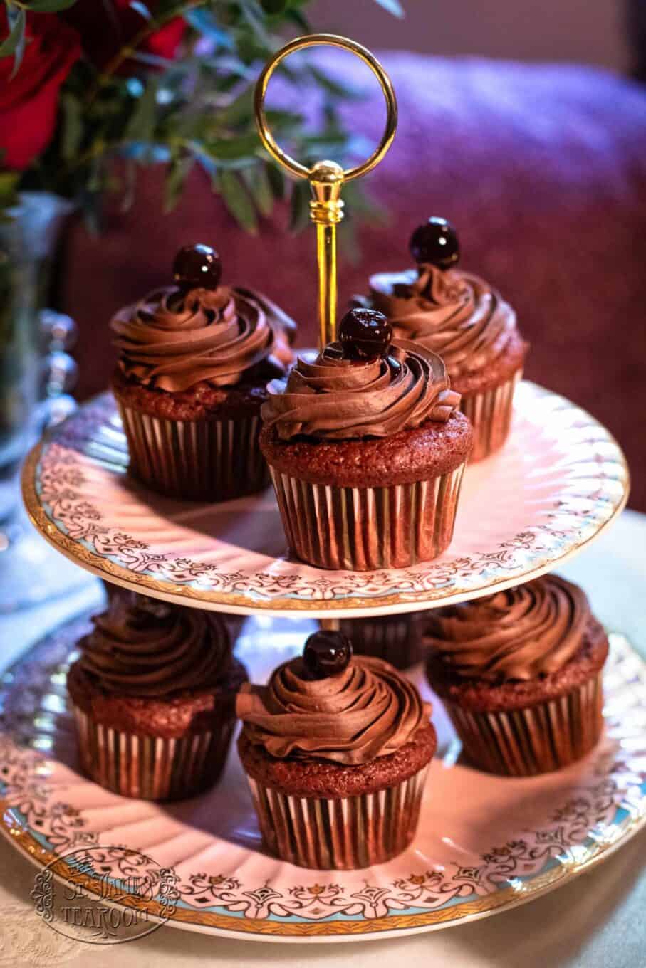 Chocolate cupcakes with swirled chocolate frosting and a cherry on top are displayed on a two-tiered, ornate china stand with a gold handle—the perfect centerpiece for any gathering. Pair these delectable treats with your favorite iced tea recipe for an irresistible afternoon indulgence.