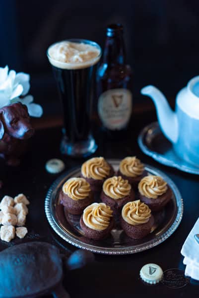 A plate of Chocolate Guinness Cupcakes with caramel frosting sits on a tray beside a pint of dark beer, a beer bottle, a white teapot, and sugar cubes, with soft lighting and decorative elements in the background.