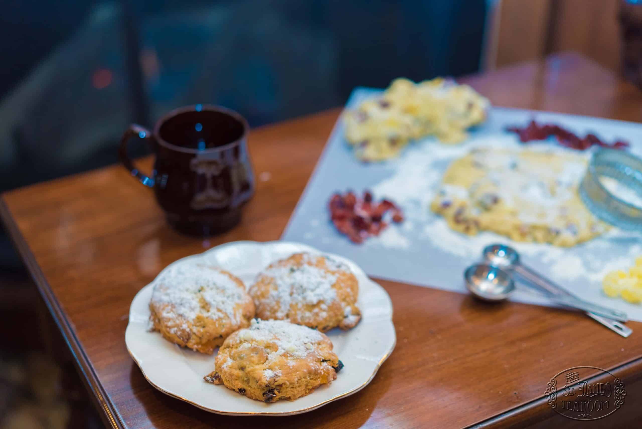 Three Christmas Stollen Scones, dusted with powdered sugar, rest on a white plate beside a dark mug on a wooden table. In the background, baking ingredients, dough, and measuring spoons are scattered across a floured surface.