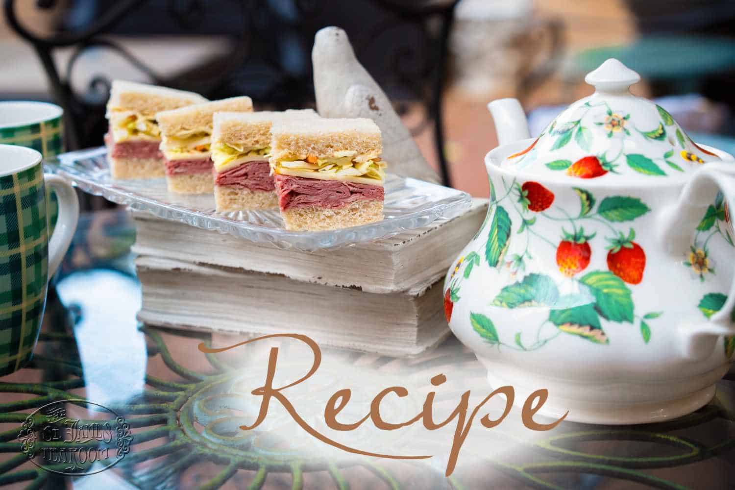 Three small tea sandwiches on a glass tray beside a strawberry-patterned teapot on a reflective table, with “Recipe” written across the image, perfectly capturing the charm of a classic Tea Sandwich Recipe.