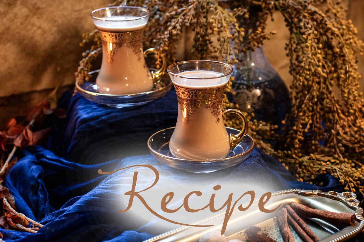 Two ornate glasses of a creamy beverage sit on saucers atop blue fabric, with dried floral decor in the background. The word “Iced Tea Recipe” is written in large script across the front.
