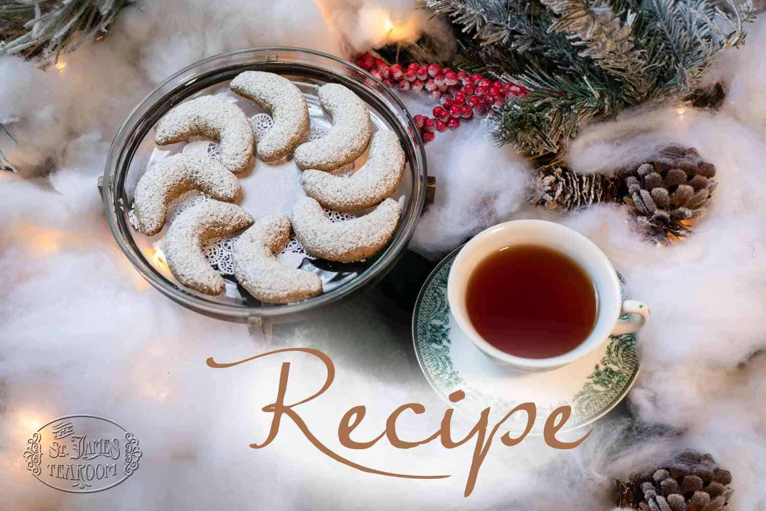 A plate of crescent-shaped cookies dusted with powdered sugar next to a cup of tea, surrounded by pinecones, faux snow, and holiday decorations. The word “Christmas Cookies Recipe” is written across the image.