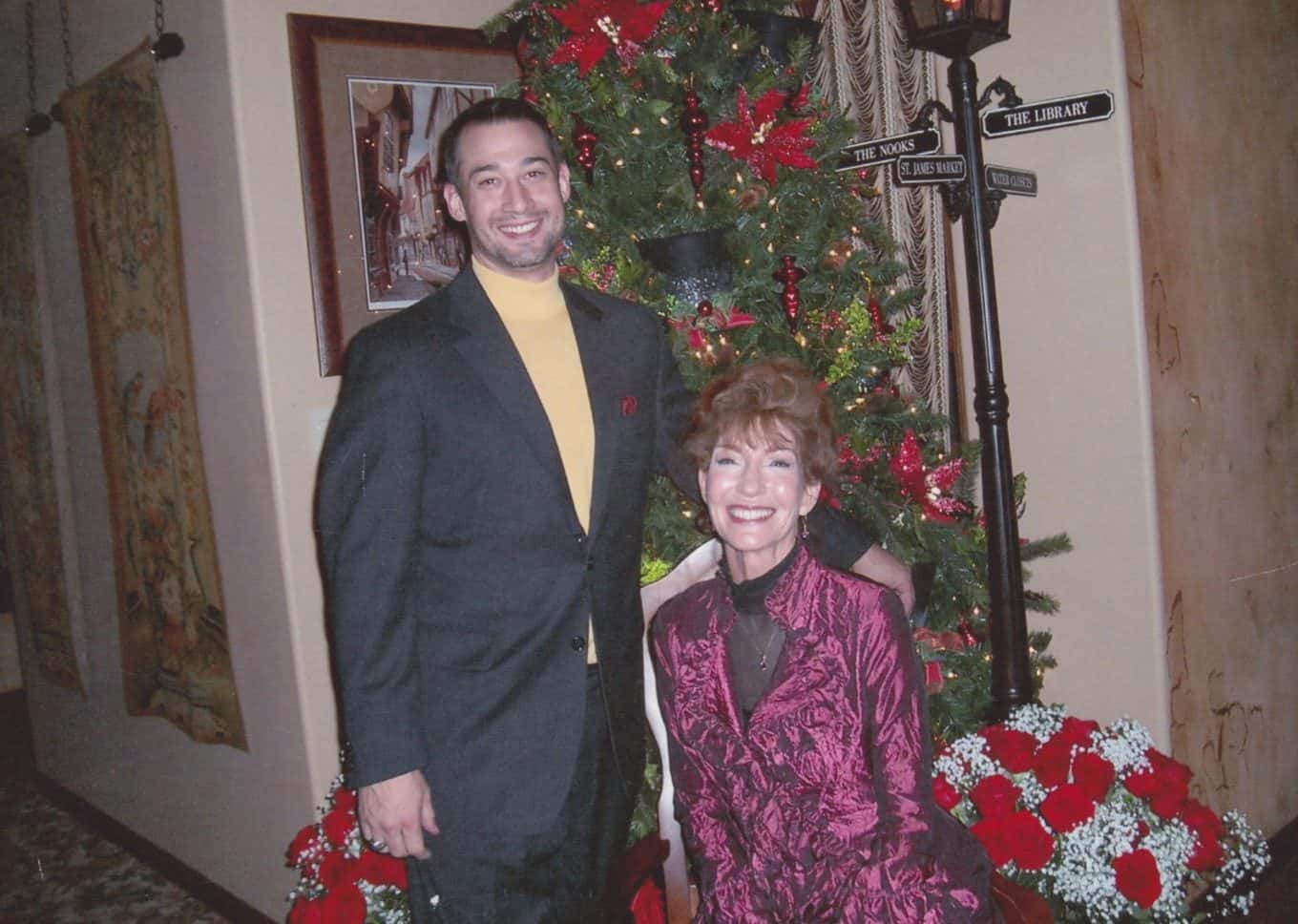 A man in a dark suit stands beside a seated woman in a red patterned jacket. They smile in front of a decorated Christmas tree with red ornaments and festive floral arrangements.
