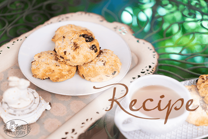 A plate of Wonderland Scones with raisins sits on a decorative tray next to a cup of tea on a green metal table outdoors. The word “Recipe” is written in large script on the image.