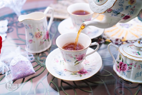 A floral teapot pours tea into a matching teacup on a glass table set with more tea cups, a creamer, and sugar bowl, all with floral patterns.