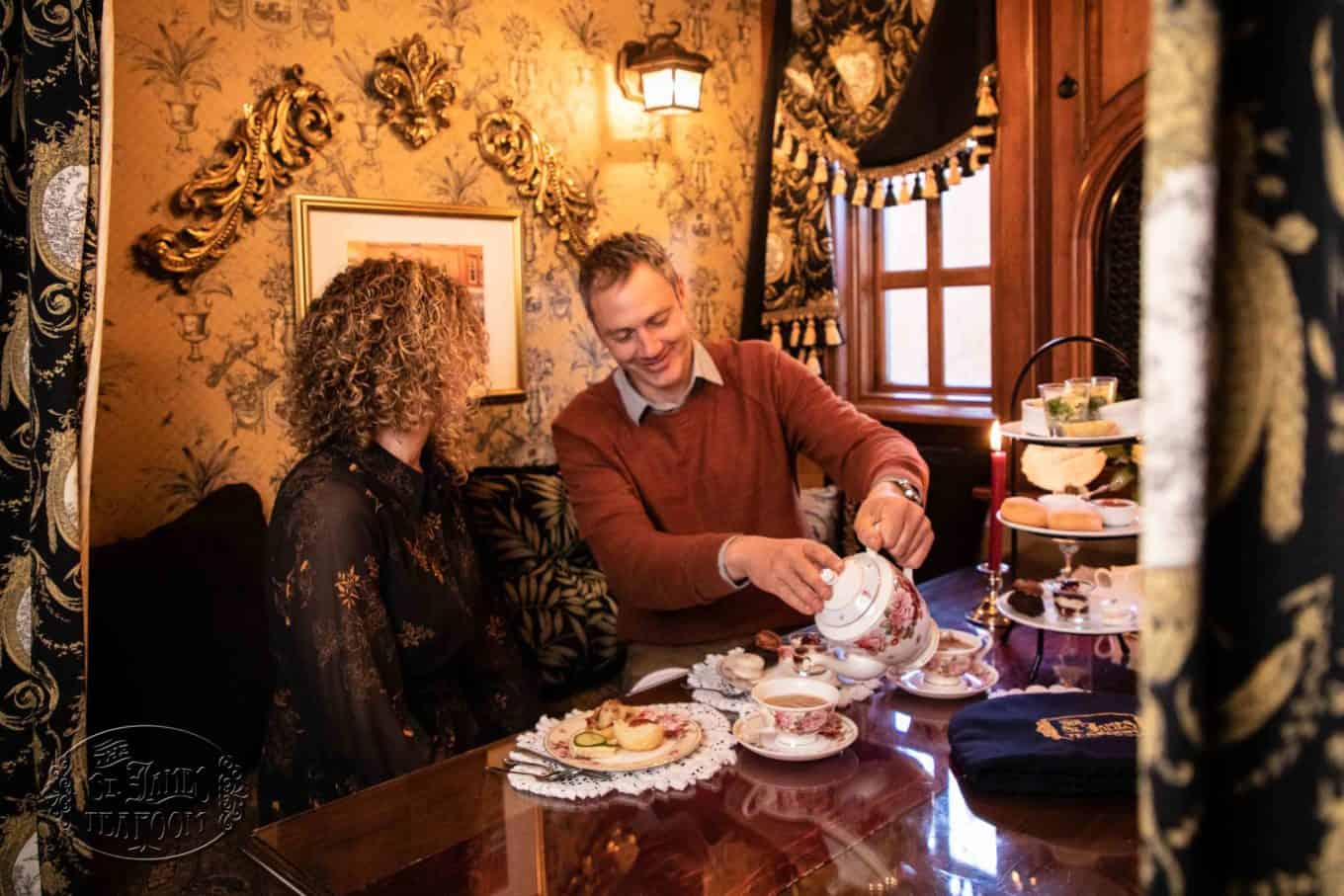 A man pours tea for a woman at an elegant table set with desserts and fine china in a richly decorated room with ornate wallpaper and golden accents, creating the perfect romantic tea room for two.