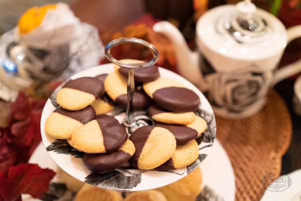 A tiered tray holds French Butter Cookies half-dipped in chocolate, next to a teapot and autumn-themed decor.
