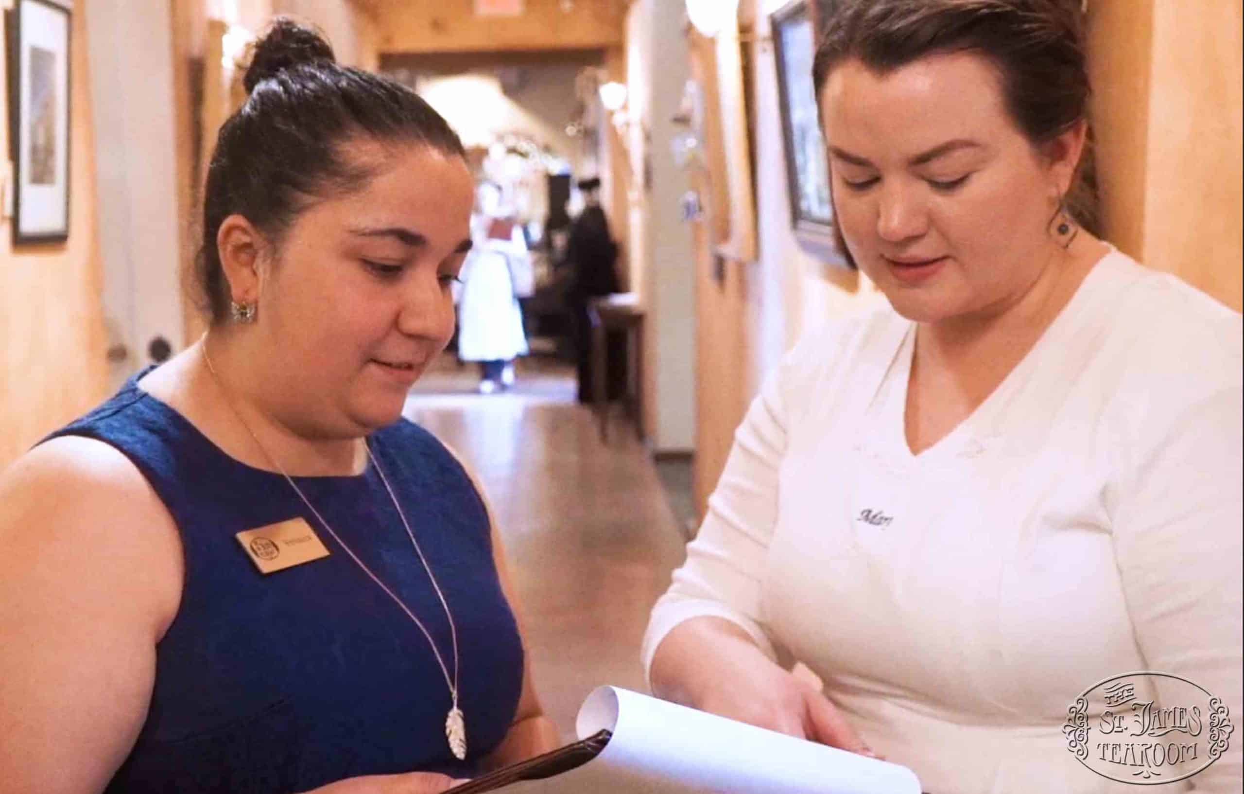 Two women stand in a hallway, one in a blue dress with a name tag and the other in white, looking at a document together. The hallway is softly lit, with framed pictures on the wall and people in the background.