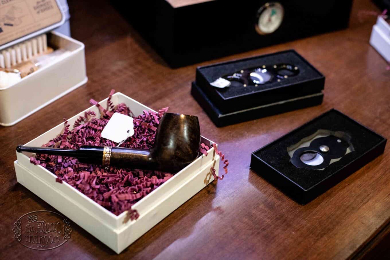A wooden smoking pipe in a gift box with decorative shredded paper sits on a table next to two black boxes containing cigar accessories.