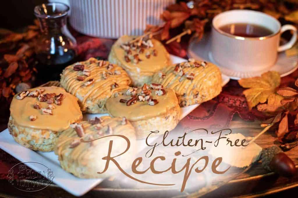 A plate of glazed scones, made using the Gluten-Free Pecan Scones Recipe with Molasses Glaze and topped with chopped nuts, sits beside a cup of tea, autumn leaves, and a small syrup jar. Text on the image reads “Gluten-Free Recipe.”.