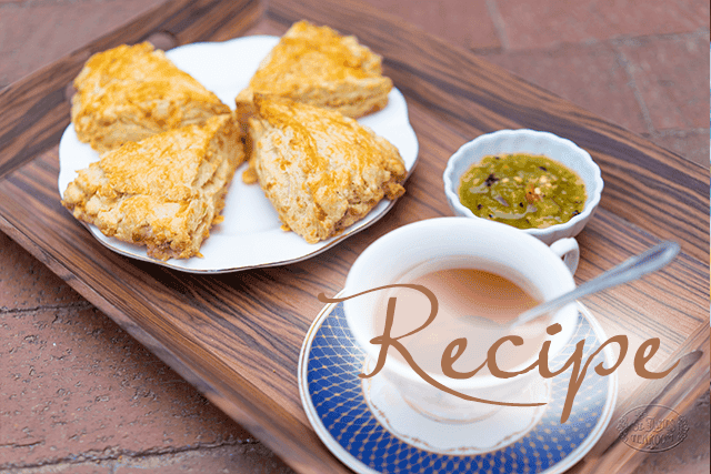 A tray with four green chile cheese scones on a white plate, a small bowl of green jam, and a cup of tea with a spoon. The word “Recipe” is written over the image.