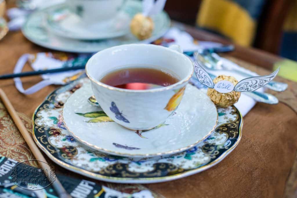 A teacup filled with tea sits on a decorative saucer, next to a Ferrero Rocher chocolate with paper wings, creating the perfect Harry Potter Teatime with your Children on a magical table setting.