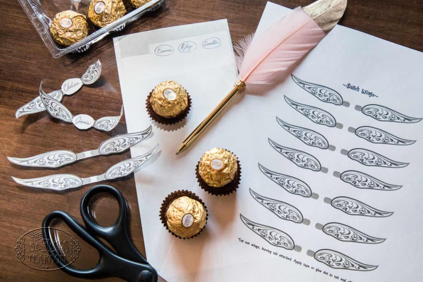 Harry Potter-themed craft scene with Ferrero Rocher chocolates, wing cutouts, scissors, a pink feather pen, and printed labels on a wooden table, showing a DIY Golden Snitch project in progress—perfect for creating magical memories during Harry Potter Teatime with your Children.