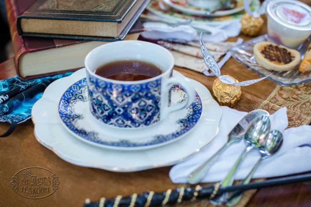 A blue and white teacup filled with tea sits on a saucer beside stacked books, wrapped chocolates, a wand, and silver spoons—creating a magical Harry Potter Teatime with your Children, all arranged on a decorative table setting.