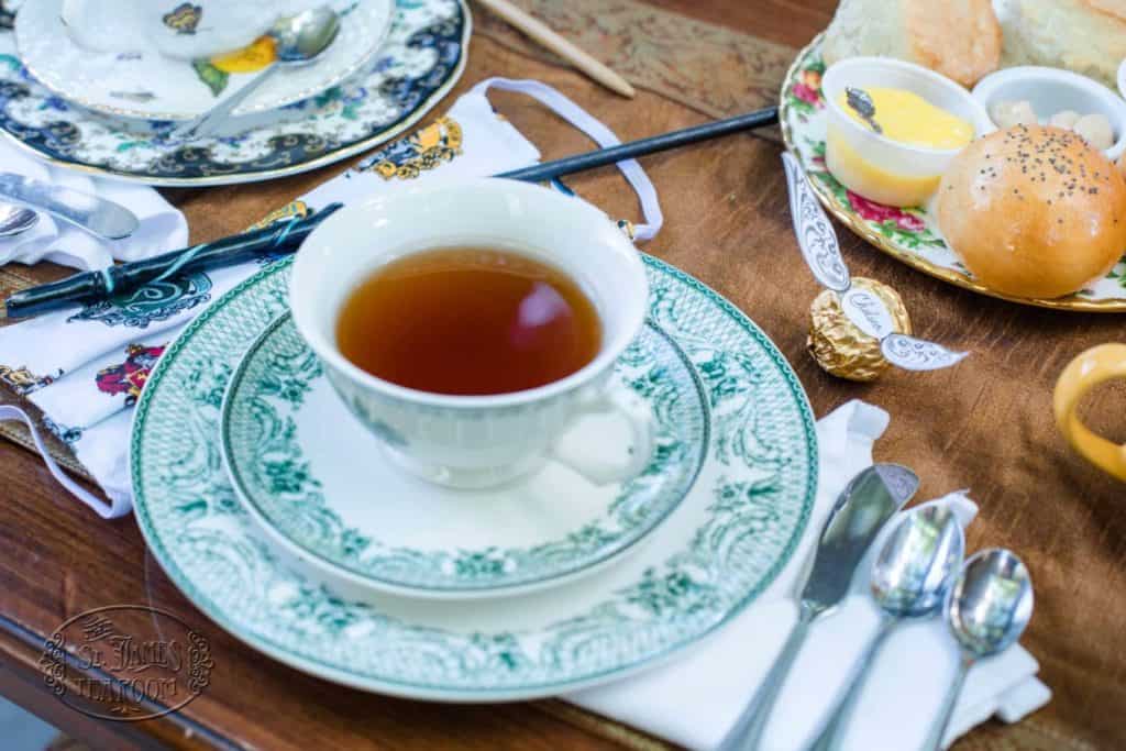 A white teacup with tea sits on a green-patterned saucer and plate, inviting you to enjoy a magical Harry Potter teatime with your children. Nearby are a buttered roll, silverware, napkin, and fancy china on a wooden table set for tea.