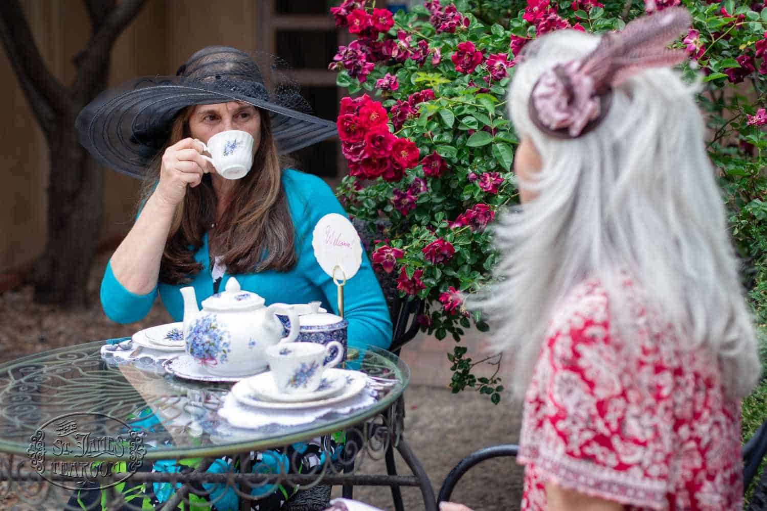 Two women in fancy hats have tea outdoors beside a blooming rose bush, using floral-patterned teapots and cups. As they savor their tea, they chat warmly about caring for fine china—one woman in blue drinks from her cup while the other, in red, faces her and listens.
