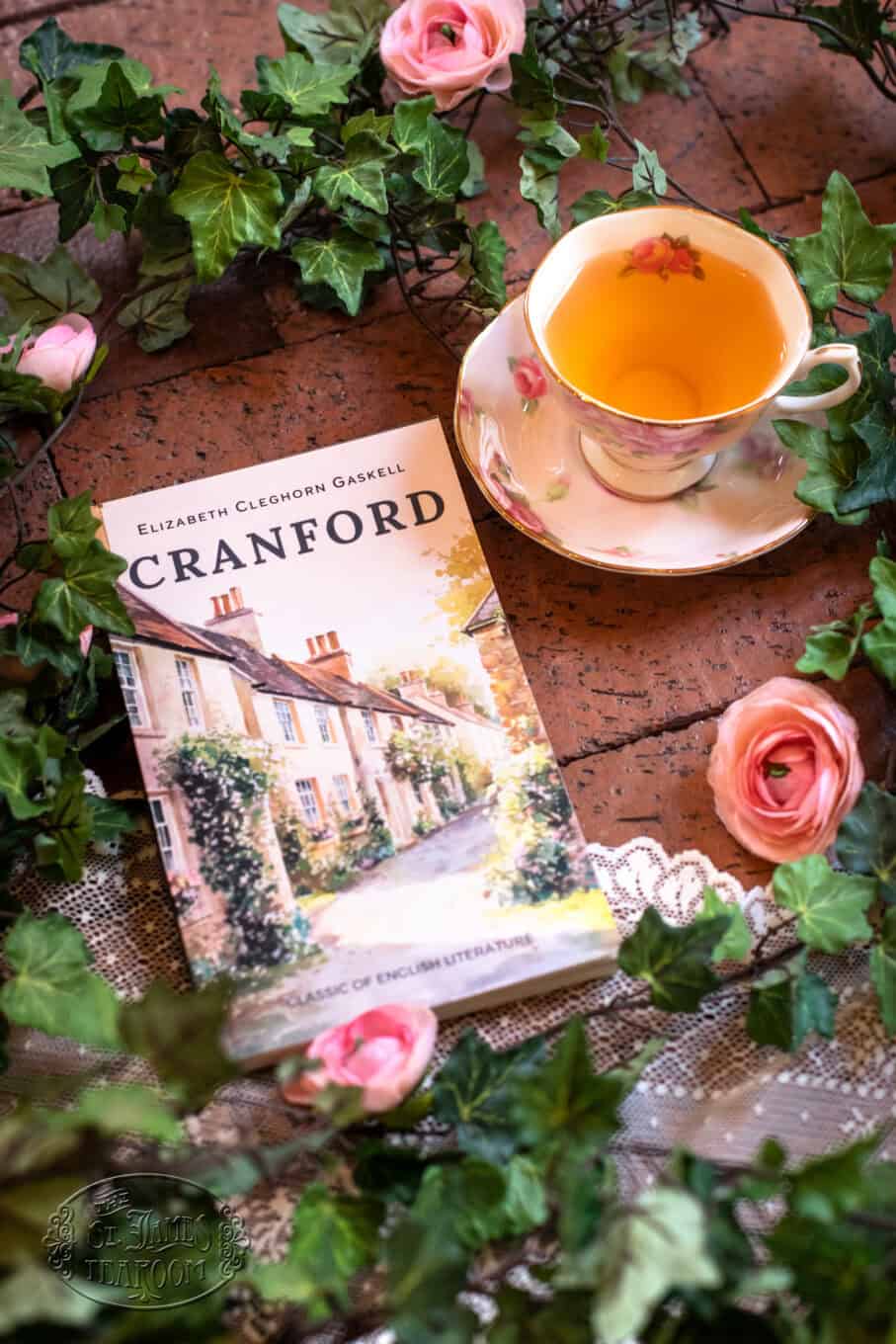 A copy of Cranford by Elizabeth Gaskell sits on a table with a floral teacup of tea, surrounded by pink artificial roses and green ivy, creating a cozy, vintage atmosphere.
