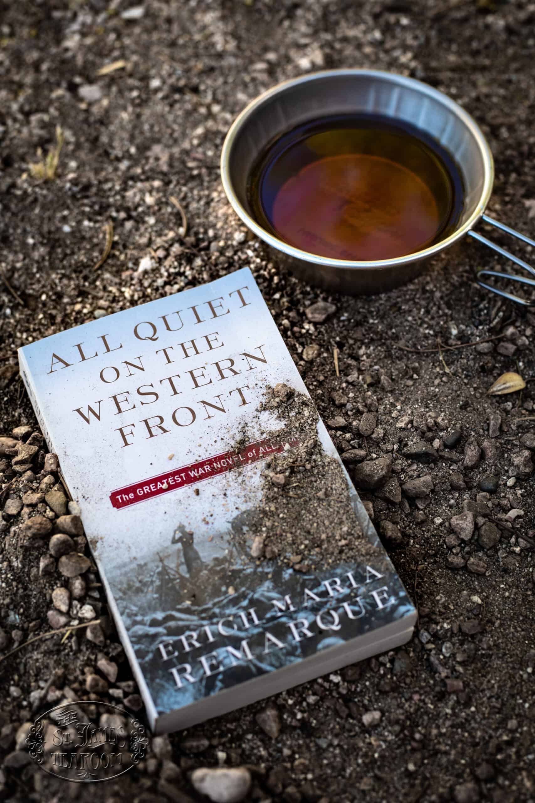 A dirt-streaked copy of “All Quiet on the Western Front” lies on rocky ground next to a metal cup filled with dark liquid.