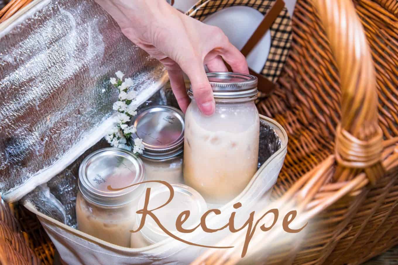 A hand places a mason jar with a silky iced tea into an insulated picnic basket. Two more similar jars and small white flowers are inside. The word “Recipe” is overlaid on the image.