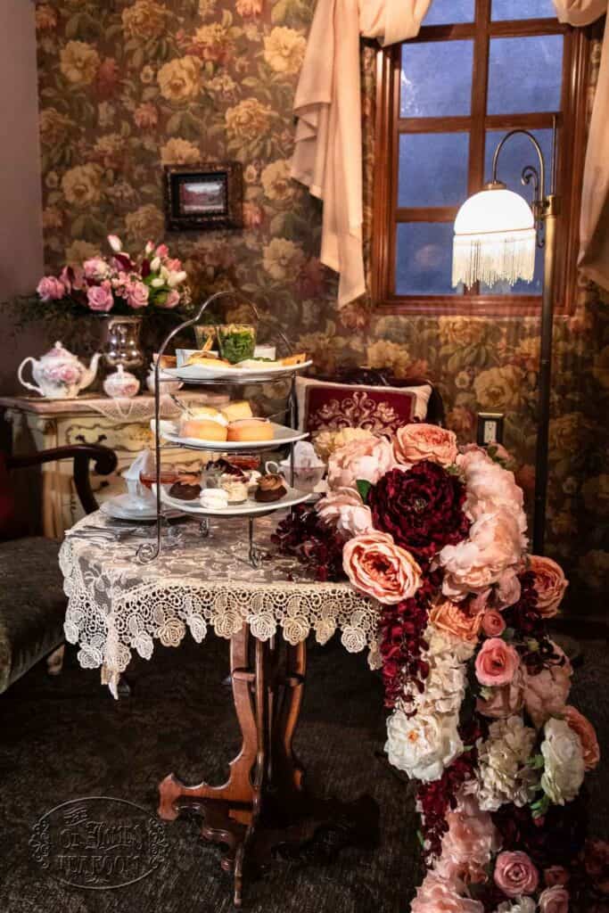 A vintage room with floral wallpaper, a lace-covered table set for tea, a three-tiered tray of desserts, and a large arrangement of pink, red, and white flowers cascading off the table.