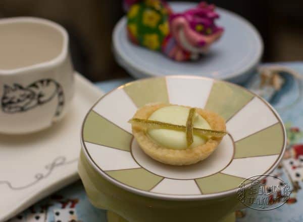 A small Lime Mousse Tart with a white filling and green garnish sits on a decorative plate next to a cup with a cat illustration, set against a blurred background featuring colorful objects.