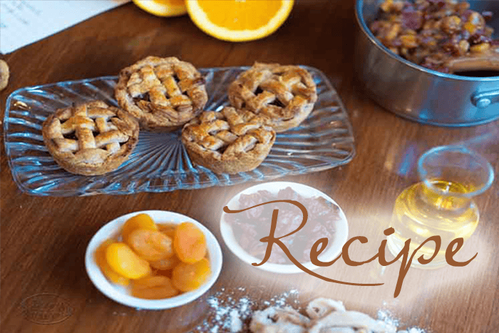 Mini Mincemeat Pies arranged on a glass plate, surrounded by dried apricots, syrup, dough, a pot with filling, and orange slices. The word “Recipe” is written across the image in elegant script.