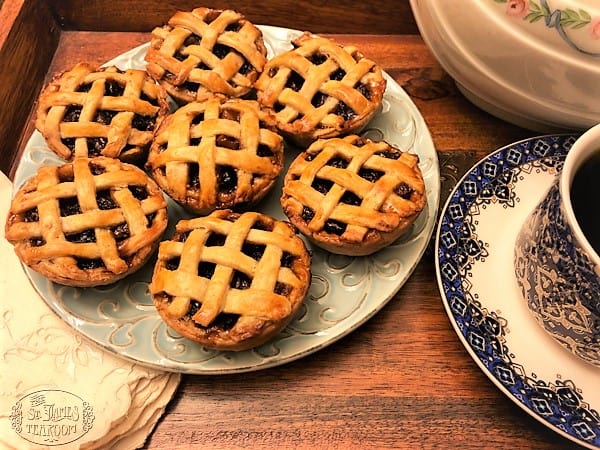 A plate of seven Mini Mincemeat Pies with lattice tops sits on a wooden tray next to a cup of black coffee in a blue-and-white patterned cup and saucer.