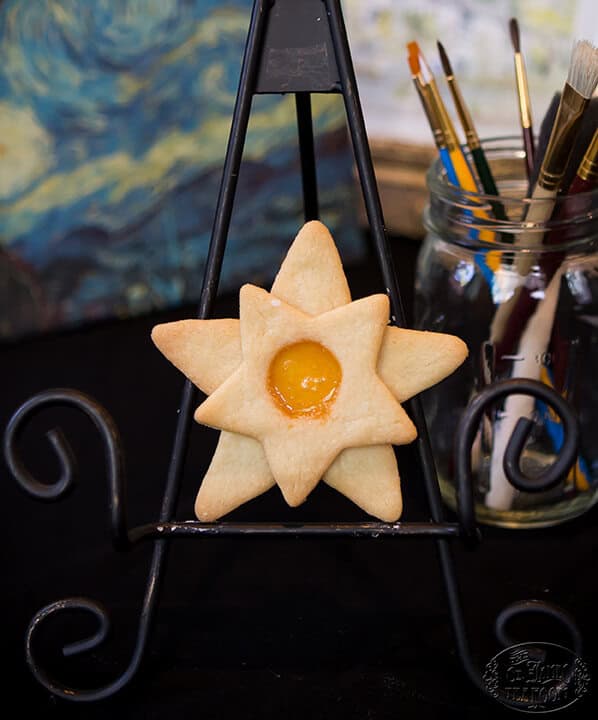 Star-shaped cookie with a yellow center displayed on a black metal stand; a jar of paintbrushes is in the background.