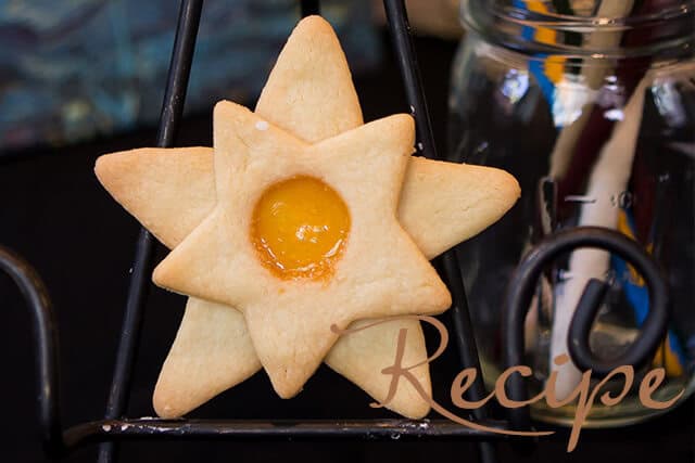 A star-shaped cookie with a yellow jam center is displayed on a stand. A glass jar and a pen are in the blurred background. The word “Recipe” is written in the lower right corner.