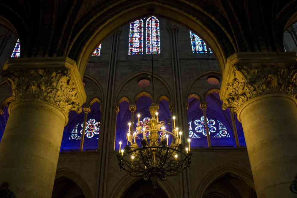 A chandelier hangs from the ceiling of a cathedral, framed by ornate columns. Like caring for fine china, every detail in this sacred space is preserved with reverence—stained glass windows glow with colorful light in the background.