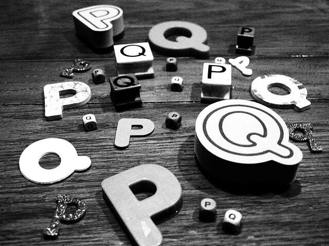 Various alphabet letters, primarily “P” and “Q” in different sizes and styles, are scattered on a wooden surface. The image is in black and white, and the letters appear to be made from different materials.