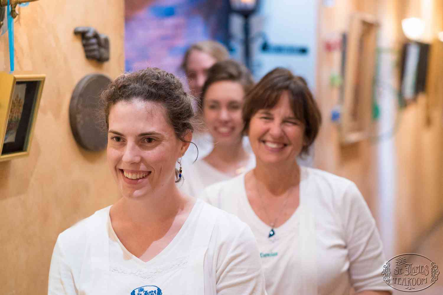 Four women stand in a line indoors, smiling and wearing white shirts. The background features framed pictures and a yellow wall. The focus is on the woman in front.