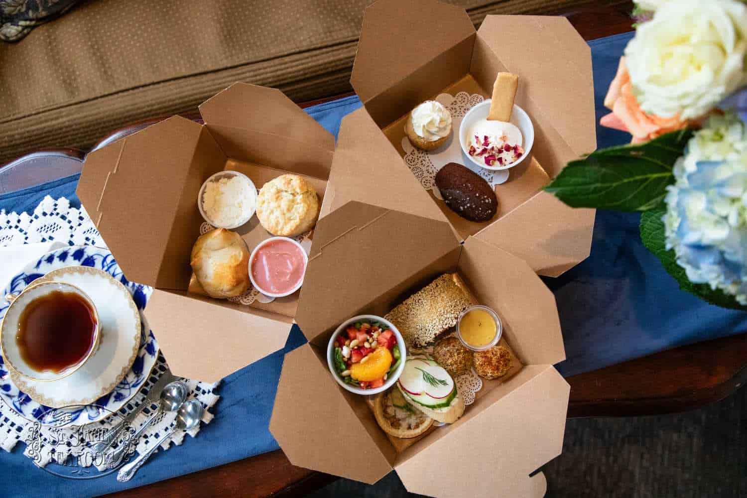 Three open cardboard boxes filled with assorted pastries, scones, fruit, and small desserts sit on a table next to a cup of tea and a vase of flowers.