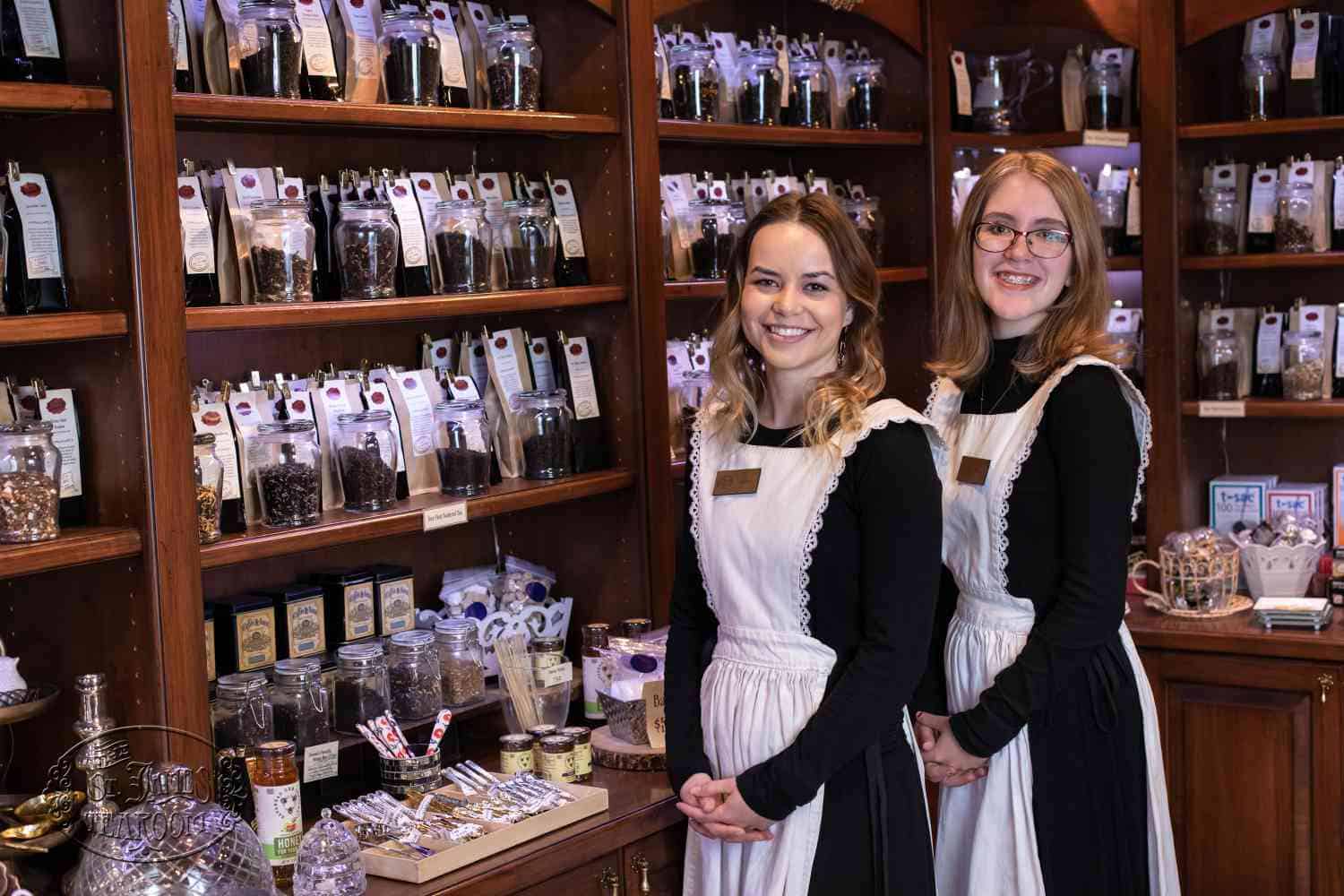 Two women wearing black tops and white aprons stand smiling in front of shelves filled with packaged goods and jars in a cozy shop. The shelves are wooden, and various candies and treats are displayed on the counter.
