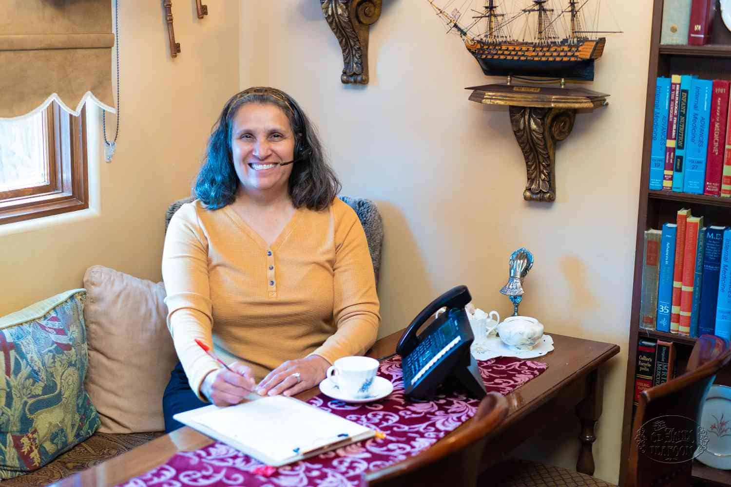 A woman with a headset smiles while writing on a clipboard at a table with a phone, teacup, and books. Model ship decor and shelves with books are visible in the cozy, warmly lit room.
