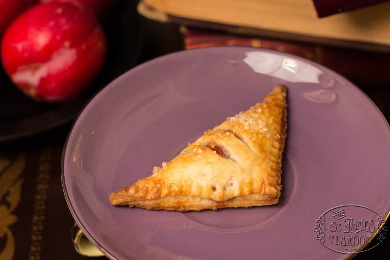 A golden-brown, triangle-shaped pastry—reminiscent of classic plum turnovers—with sugar crystals sits on a purple plate. Red apples are blurred in the background, and the plate features a faint “St. James Tearoom” logo.